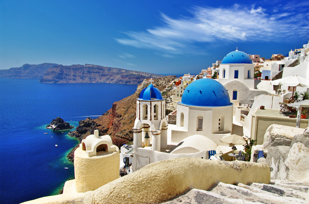 Blick auf die weiße Architektur der Stadt Santorini mit ihren charakteristischen blauen Kuppeln, die an einer steilen Klippe über dem tiefblauen Meer liegen. Im Hintergrund ist eine felsige Insel im Wasser zu sehen, der Himmel ist klar mit wenigen weißen Wolken.